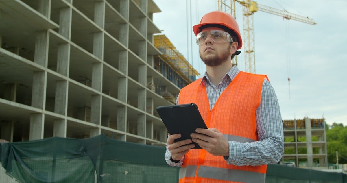 A young architect or builder standing on a construction site, holding a digital touchpad and reviewing plans with structures in the background.